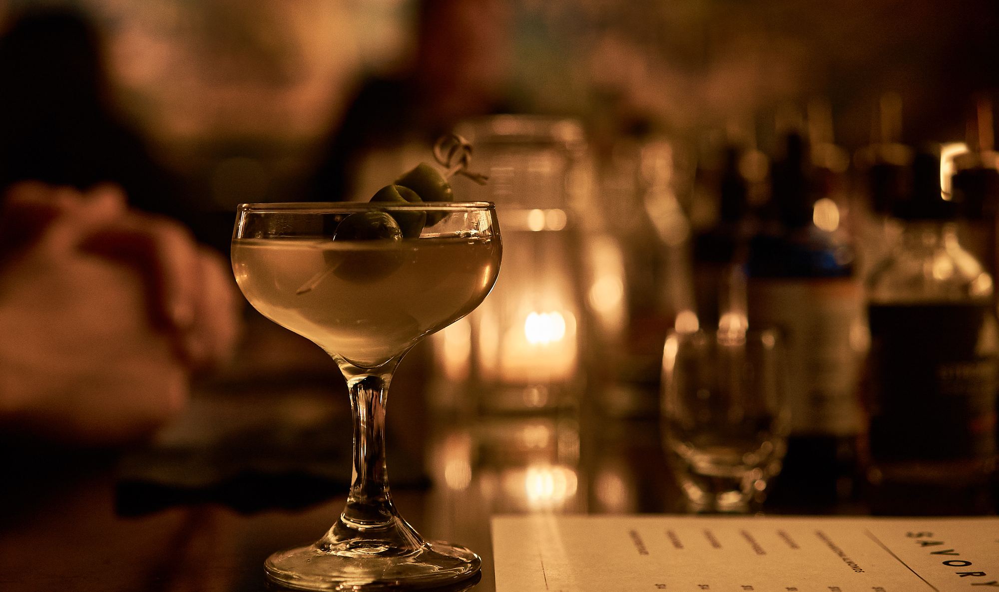 A martini garnished with olives on the bar, lit by candlelight
